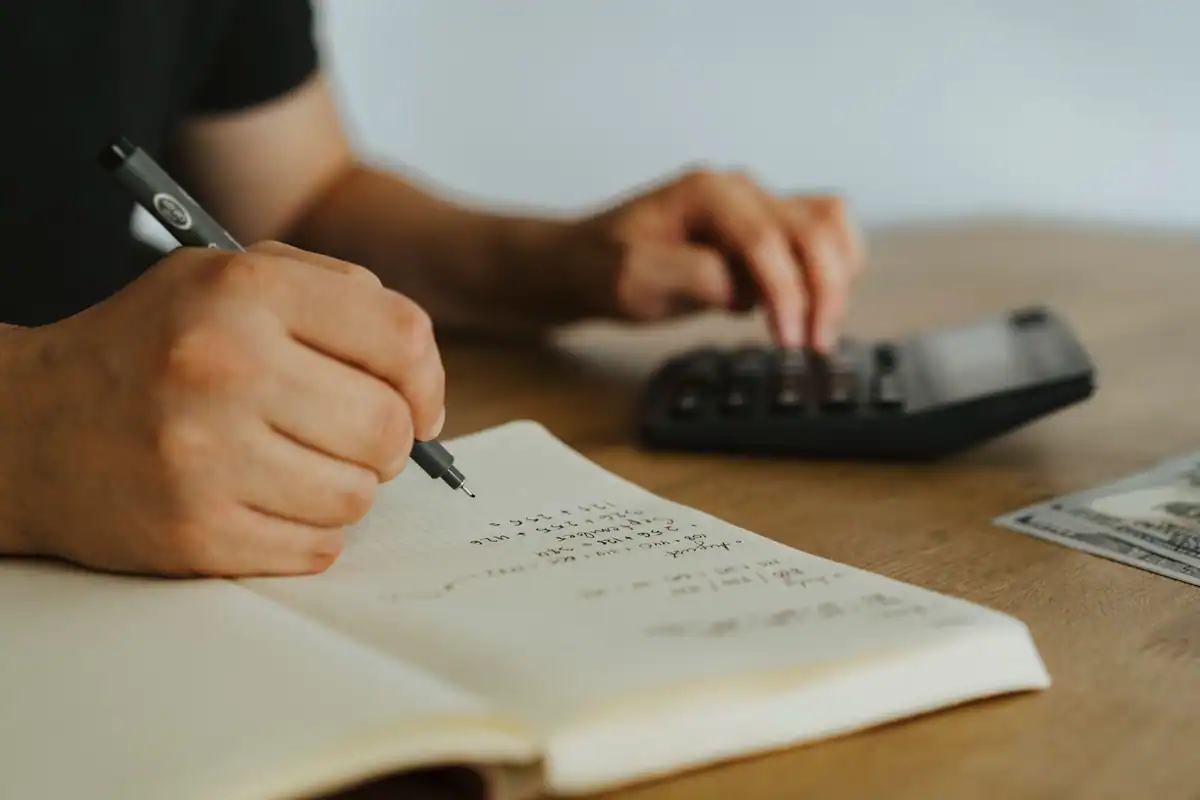 Mâini care notează calcule într-un caiet, lângă un calculator, pentru bugetul mutării
